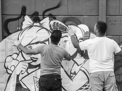 A black and white photograph of two men pressing large printed paper sheets onto a wall, the outline of two girls, one with a bow in her hair, drawn across the paper