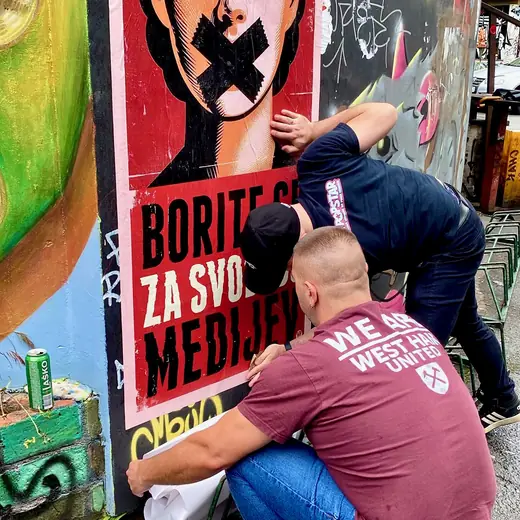 Two people pasting the poster: a woman with her mouth taped shut over the words Borite se za svobodo medijev