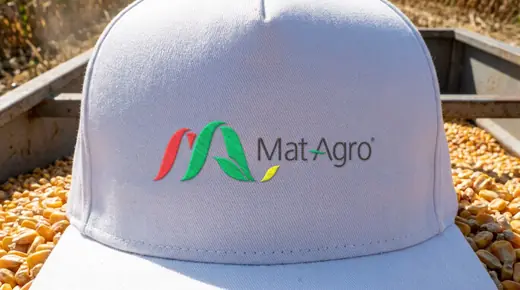 MatAgro: the horizontal Mat Agro logo embroidered in red, green, yellow and grey satin stitches on the front of a white cap resting on shelled corn in a grain trailer, close up, sunlight raking across the threads