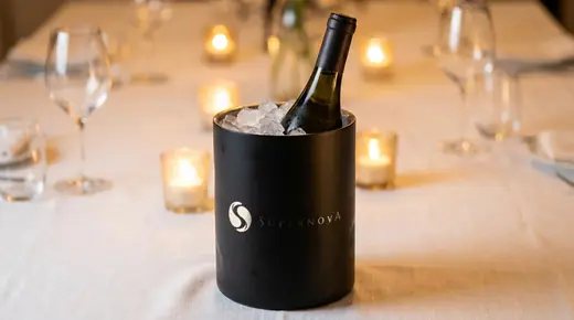 Supernova: the white simplified Supernova logo, the S of two drops and the SUPERNOVA wordmark, printed on a matte black ice bucket holding a bottle of white wine on a linen tablecloth at a candlelit wine tasting, glasses blurred behind
